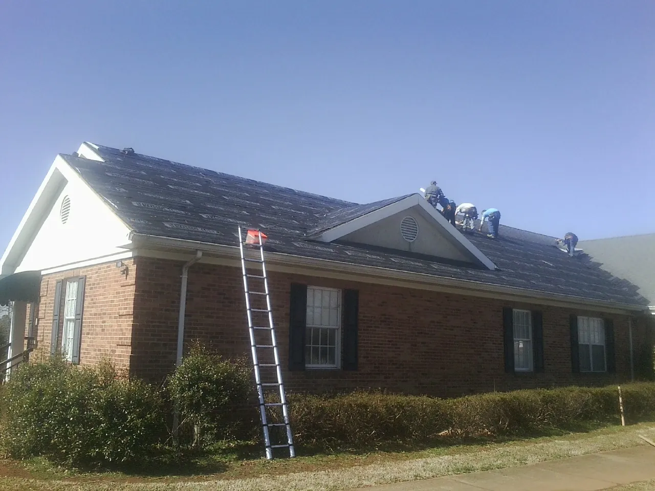 Crew tearing shingles off a brick ranch roof with a ladder set — Metal Roof Repair work by our crew in Summit Point