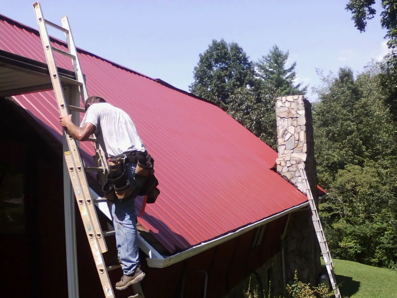 Roofer on a ladder beside a red metal roof and stone chimney — Soffit & Fascia Repair by Roofing Brothers in Summit Point