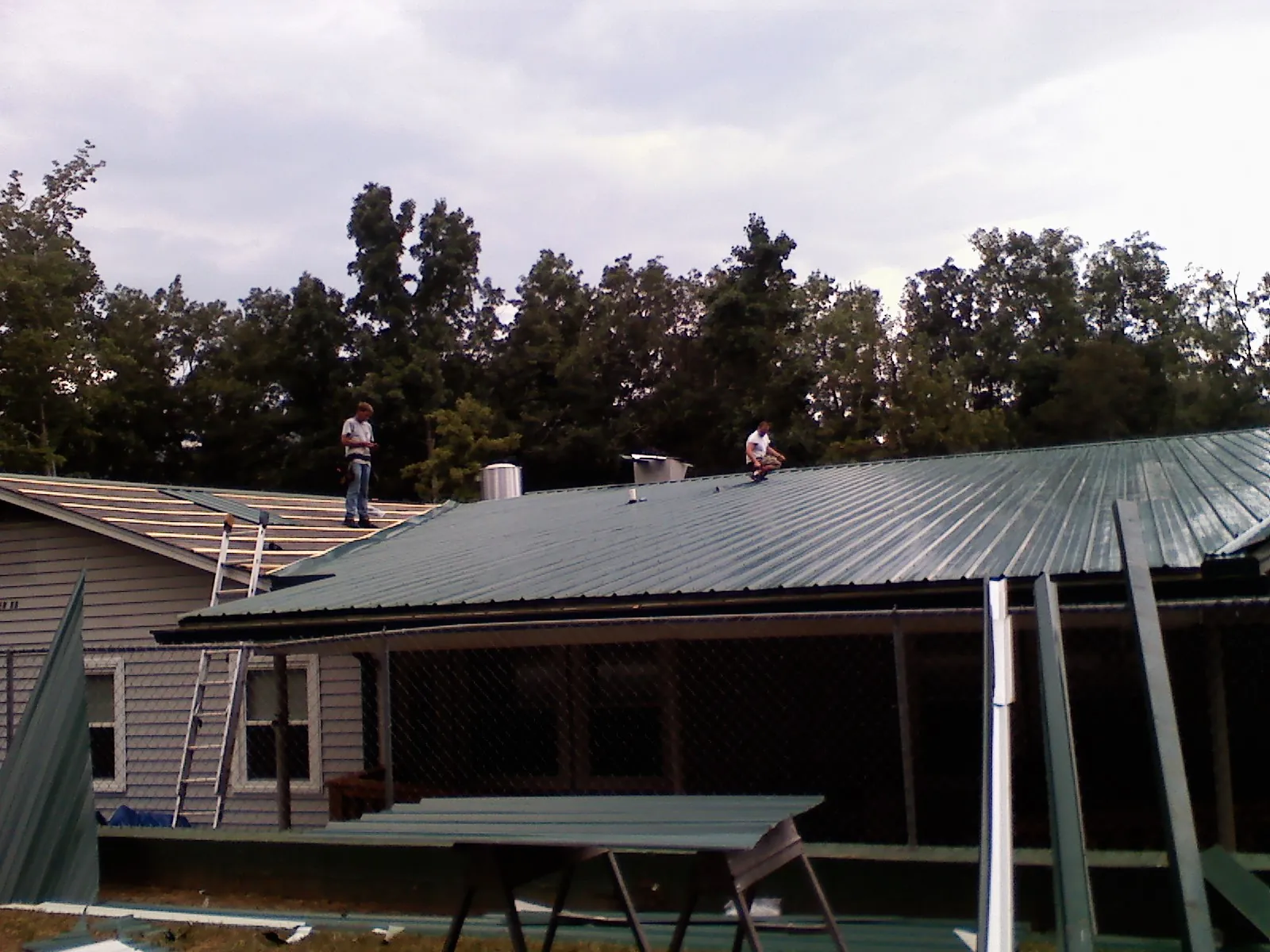 Two roofers installing green metal panels above a covered porch in Summit Point